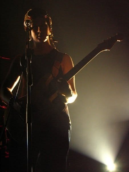Shaamans on stage: a guitarist silhouetted against a white spotlight, playing an electric guitar at a microphone stand.